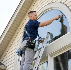 Professional window cleaner on a ladder using a squeegee to clean an exterior arched window, San Jose, CA.
